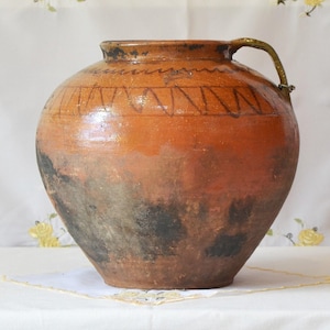 May include: A large, round, antique earthenware jug with a handle. The jug is primarily brown with darker areas near the base and a decorative band of zigzags near the top. The handle is a lighter color.