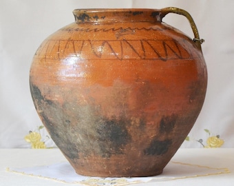 Op de afbeelding: Een grote, ronde, antieke aardewerken kruik met een handvat. De kruik is voornamelijk bruin met donkere plekken bij de basis en een decoratieve band van zigzags nabij de bovenkant. Het handvat heeft een lichtere kleur.