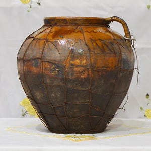 May include: A large, antique ceramic jar with a handle, featuring a dark brown and tan color scheme. The jar is encased in a wire mesh, adding a rustic aesthetic. The jar's surface has a glossy finish, and it sits on a white surface.