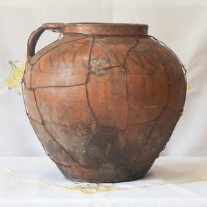 Puede incluir: Una jarra grande y antigua de terracota con un asa. La jarra está envuelta en una malla de alambre para soporte. El recipiente de cerámica tiene un aspecto rústico y envejecido con variaciones en tonos marrones y desgaste visible.