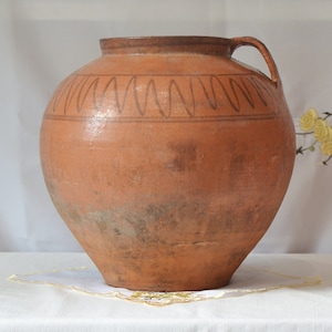 May include: A large, brown, earthenware pot with a wide mouth and a single handle. The pot has a decorative pattern of wavy lines around the top.