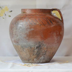May include: A large, brown, earthenware pot with two handles. The pot has a rough, textured surface and appears to be very old.