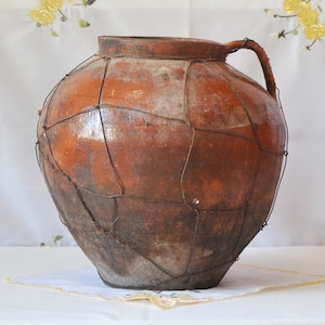 May include: A large, brown clay pot with a wire mesh covering. The pot has a wide mouth and a handle on one side.