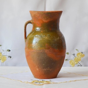 Può includere: Brocca rustica in ceramica con manico. Presenta una miscela di toni terrosi, tra cui arancione, marrone e verde. La brocca, probabilmente fatta a mano, è destinata a contenere liquidi e ha un'ampia apertura e un manico curvo.