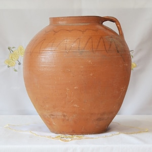 May include: A large, terracotta-coloured clay pot with a wide mouth and a single handle. The pot has a decorative design of wavy lines around the top.