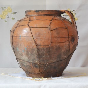 Puede incluir: Una olla de barro grande de color terracota con una cubierta de malla de alambre. La olla tiene una boca ancha y un asa en un lado. La olla está sentada sobre un mantel blanco.