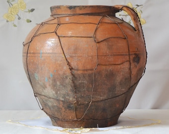 Op de afbeelding: Een grote, terracottakleurige kleipot met een gaas van draad eromheen. De pot heeft een brede opening en een handvat aan één kant. De pot staat op een wit tafelkleed.