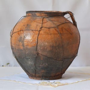 May include: A large, antique terracotta pot with a wire cage. The pot has a rounded body, a handle, and a dark, aged appearance. The wire cage is wrapped around the pot, adding a rustic, vintage aesthetic. The pot is likely used for storage or decoration.
