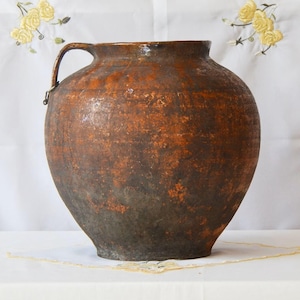 Peut inclure: Une grande cruche en terre cuite de style antique avec une anse. La cruche a un corps arrondi et une large ouverture. L'extérieur est un mélange moucheté de teintes marron et orange, lui donnant un aspect rustique. La cruche repose sur une surface blanche.