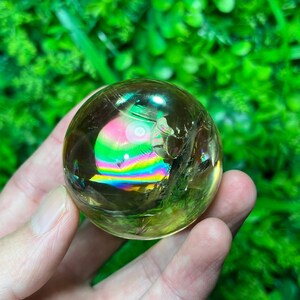 May include: A clear, spherical crystal ball with internal rainbow refractions. The ball is held in a hand against a backdrop of green foliage. The crystal has a golden hue and displays a spectrum of colors.