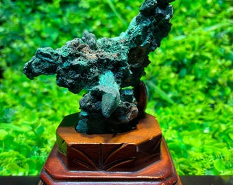 May include: A green mineral specimen on a wooden display stand. The mineral has a rough, textured surface and appears to be a type of malachite.