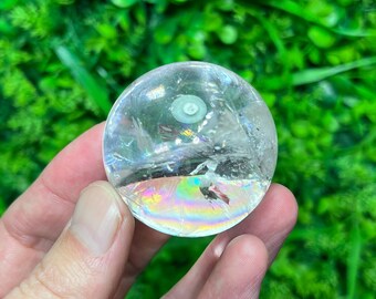 May include: A transparent crystal sphere with internal fractures and a rainbow sheen. The ball features a small, pale green inclusion at the top. Set against a vivid green backdrop, the sphere stands out.