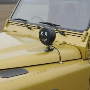 Può includere: Un faro rotondo nero con un design a faccina sorridente è montato sul cofano di un veicolo giallo. La luce ha due segni 'X' bianchi per gli occhi e una bocca sorridente bianca. Sono visibili anche i tergicristalli.
