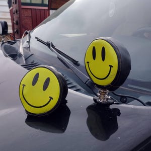 Op de afbeelding: Twee gele smiley-gezichtshoezen met zwarte rand, één bevestigd aan de koplamp van een voertuig en de andere rustend op de motorkap. De hoezen hebben een klassiek smiley-gezichtsontwerp met zwarte ogen en een gebogen mond.