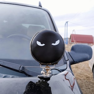 May include: A black, round car accessory with white, stylized eyes, mounted on the hood of a vehicle. The accessory is made of a leather-like material and has a metal mounting bracket. The background shows a cloudy sky and a rural setting.