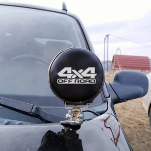 Op de afbeelding: Een ronde zwarte offroad-lamp met een wit "4x4 OFF ROAD"-logo gemonteerd op een zwarte auto.