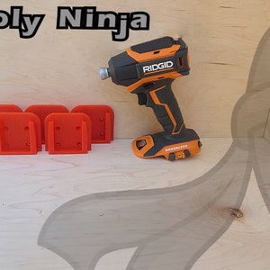 Pu&ograve; includere: Quattro supporti a parete arancioni stampati in 3D per un trapano, accanto a un trapano Ridgid nero e arancione su una superficie in legno. Lo sfondo &egrave; un viso di ninja dei cartoni animati.