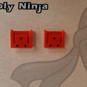 May include: Two bright orange plastic wall mounts with four black screw holes each. The mounts are attached to a light wood surface with a cartoon ninja illustration.