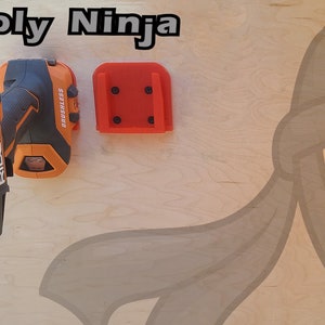 Pu&ograve; includere: Un trapano a batteria nero e arancione con una punta argentata, montato su un supporto a parete rosso stampato in 3D. Il testo "3D Supply Ninja" &egrave; visibile nella parte superiore dell'immagine.