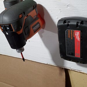 Puede incluir: Tres herramientas el&eacute;ctricas colgadas en una pared blanca. Un taladro inal&aacute;mbrico negro y naranja con una broca roja, un taladro inal&aacute;mbrico negro con un bot&oacute;n rojo y una bater&iacute;a negra con la palabra "Milwaukee" impresa en ella.
