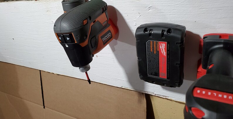 Puede incluir: Tres herramientas el&eacute;ctricas colgadas en una pared blanca. Un taladro inal&aacute;mbrico negro y naranja con una broca roja, un taladro inal&aacute;mbrico negro con un bot&oacute;n rojo y una bater&iacute;a negra con la palabra "Milwaukee" impresa en ella.