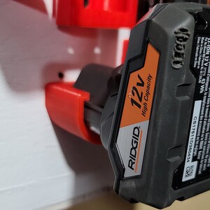 May include: A black and orange Ridgid 12V High Capacity battery with a red plastic holder. The battery has a label with the text "RB2059", "WARNING/Avertissement", "LI-ION 12V", "43 Wh", "C51761110360434", "12v", "RIDGID", "High Capacity".