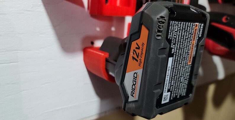 May include: A black and orange Ridgid 12V High Capacity battery with a red plastic holder. The battery has a label with the text "RB2059", "WARNING/Avertissement", "LI-ION 12V", "43 Wh", "C51761110360434", "12v", "RIDGID", "High Capacity".