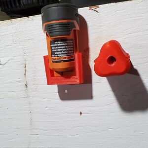 Puede incluir: Un soporte para bater&iacute;as impreso en 3D de color naranja y negro con una base roja y un gancho rojo impreso en 3D. El soporte para bater&iacute;as est&aacute; montado en una pared blanca.