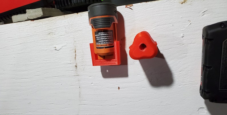 Puede incluir: Un soporte para bater&iacute;as impreso en 3D de color naranja y negro con una base roja y un gancho rojo impreso en 3D. El soporte para bater&iacute;as est&aacute; montado en una pared blanca.