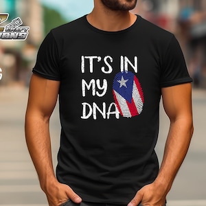 Puede incluir: Una camiseta negra con el texto "It's in my DNA" en blanco con un aspecto desgastado. Un gráfico de huella dactilar con los colores de la bandera puertorriqueña está al lado del texto.