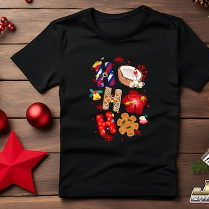 Puede incluir: Camiseta negra con un diseño colorido de "Ho Ho" que incluye un hombre de jengibre, un coco, una flor de hibisco y una máscara tiki.