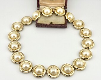 May include: A gold-tone metal necklace featuring a series of circular links, each set with a central cream-colored faux pearl. The necklace is arranged in a circular shape in front of an open, rectangular, dark-colored jewelry box with a cream lining.