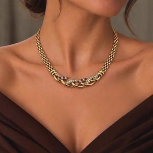May include: A gold chain necklace featuring a central braided design. The centerpiece is embellished with alternating round clear stones and round red stones set within the interlocking gold links of the collar-style necklace.