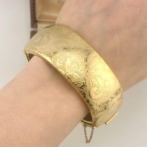 May include: A wide, gold-colored bangle bracelet with an ornate floral pattern. The hinged bracelet is secured with a clasp and a safety chain. The bracelet is approximately 2 inches wide.