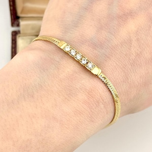 May include: A gold bracelet with a herringbone chain and a rectangular centerpiece. The centerpiece features five clear, round gemstones. The bracelet is worn on a wrist.