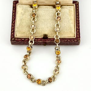 Puede incluir: Un collar de cadena de metal en tono dorado que presenta una serie de eslabones circulares, cada uno engastado con un cristal redondo facetado en varios tonos ámbar, miel y marrón claro, dispuestos en un patrón repetitivo a lo largo.