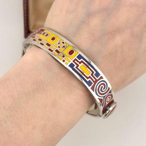 Op de afbeelding: Zilverkleurige scharnierende armband met een kleurrijk geometrisch ontwerp. De armband heeft rode, gele en blauwe emaille accenten. Het ontwerp omvat vierkanten, rechthoeken en spiraalvormen. De armband heeft een diameter van ongeveer 6,35 cm.