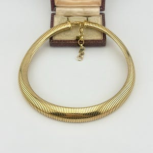 May include: A gold-toned omega chain necklace featuring a wide, semi-rigid, ribbed design that forms a circular collar shape. The necklace includes a short extender chain with small circular links attached to the back of the collar structure.