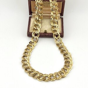 Puede incluir: Un collar de cadena en tono dorado con un diseño de doble eslabón y superficies texturizadas en cada eslabón. El collar está drapeado en forma de U, con los extremos superiores descansando dentro de un joyero rectangular de color burdeos.