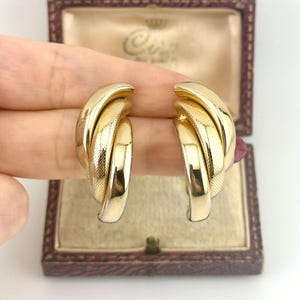 May include: A pair of gold-toned earrings with a textured, interwoven design. The earrings are in a classic hoop style, with a polished finish. The earrings are held in a vintage jewelry box with a maroon exterior and a cream-colored interior.