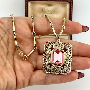 Könnte beinhalten: Eine goldfarbene Halskette mit einem rechteckigen Anhänger mit einem großen, rosafarbenen, smaragdgeschliffenen Edelstein. Der Anhänger ist von einem verzierten, durchbrochenen Design mit kleineren rosafarbenen Edelsteinen umrahmt.