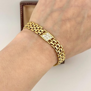 May include: A gold-toned metal link bracelet featuring a repeating brick-like pattern. The centre of the bracelet displays a square-shaped focal point embedded with nine small, clear, round-cut stones arranged in a three-by-three grid formation.