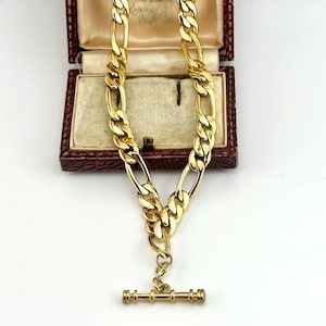 Könnte beinhalten: Eine goldfarbene Kettenhalskette mit einem sich wiederholenden Muster aus drei kleinen runden Gliedern, gefolgt von einem länglichen ovalen Glied. Die Kette endet in einem T-Steg-Verschluss, einem horizontalen Metallstab mit gerillten Enden.