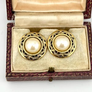 May include: A pair of round gold-tone earrings featuring a central cream-colored faux pearl cabochon surrounded by an intricate filigree metal border with a textured edge, presented inside a rectangular box with a cream velvet interior lining.