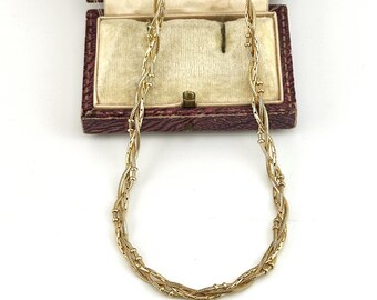 May include: A gold-toned necklace featuring a multi-strand braided design with thin, textured chains and small spherical beads. The necklace is displayed in front of an open, rectangular, burgundy-colored jewelry box with a cream-colored interior.