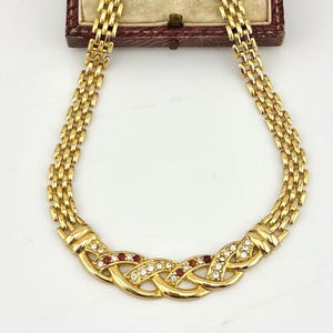 May include: Gold-coloured necklace with a braided centre design. The necklace features clear and red gemstones set within the braided section. The chain is made of small, rectangular links. The necklace is displayed on a white surface.