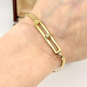 May include: A gold-toned bracelet featuring a delicate chain band connected to a central rectangular bar element. The bar contains a single, small, clear, round-cut stone set in a star-shaped metal mount that slides along the open center.
