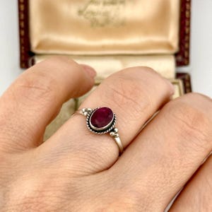 Può includere: Un anello d'argento con una gemma ovale rosso scuro. La gemma è incastonata in una lunetta decorativa con un motivo a corda intrecciata. L'anello presenta piccoli accenti di perline su entrambi i lati della gemma.