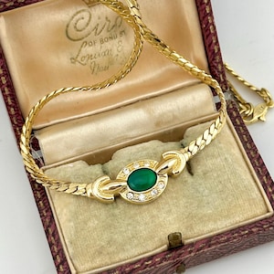 May include: A gold-toned necklace with a central oval-shaped green stone, surrounded by small clear stones. The necklace is displayed in a vintage jewelry box with a maroon exterior and a cream-colored interior.