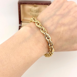Puede incluir: Una pulsera de cadena tipo cuerda en tono dorado con un diseño de eslabones entrelazados y retorcidos. La pulsera tiene una superficie pulida y reflectante, y se lleva alrededor de la muñeca, mostrando el patrón en espiral continuo de los eslabones.